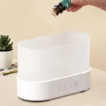 A person adding essential oil drops into a white ultrasonic diffuser on a clean surface.