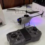 A small remote-controlled helicopter with a gray body and black rotor blades, displayed on a table. The helicopter features LED lights and is operated via a black remote control with multiple buttons.