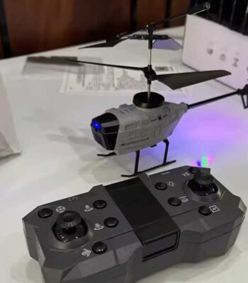 A small remote-controlled helicopter with a gray body and black rotor blades, displayed on a table. The helicopter features LED lights and is operated via a black remote control with multiple buttons.