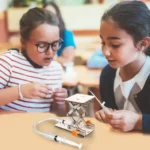Two young girls exploring a science robotics kit with a small mechanical robot on a classroom table, engaging in hands-on STEM learning activity.