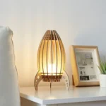 A modern wooden table lamp with a unique, artistic design, placed on a white surface next to a framed photo and a potted plant.