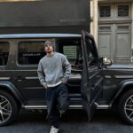 Young man leaning against a black SUV with open door, dressed in casual gray sweater and beanie, urban street background.