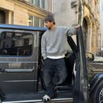 A young man in casual attire, including a gray sweater and beanie, exiting a black SUV parked on a city street with historic buildings in the background.