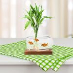 A decorative plant in a glass fishbowl with goldfish and green leaves, placed on a wooden base on a checkered green and white cloth.