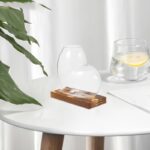 A modern glass vase with a unique, rounded shape on a wooden base, placed on a white table with a glass of lemon water and a plant in the background.