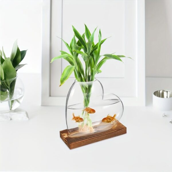Green plant in a glass fishbowl with three goldfish, placed on a wooden base, on a white surface with a white background.