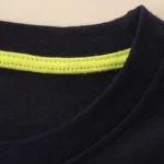 Close-up of a black T-shirt neckline featuring neon yellow stitching detail, highlighting the fabric texture and seam quality.