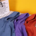 Blue, purple, and red hoodies with drawstrings, laid out on a grid surface against a yellow backdrop.